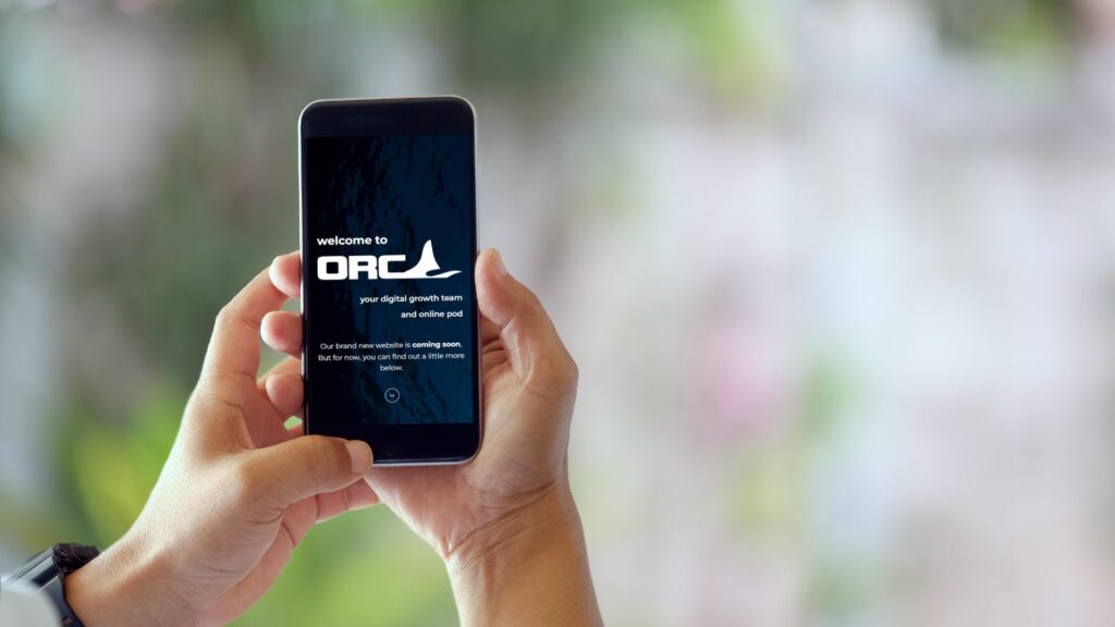 A pair of hands holding a mobile phone with the Orca Online Marketing website homepage on the screen.